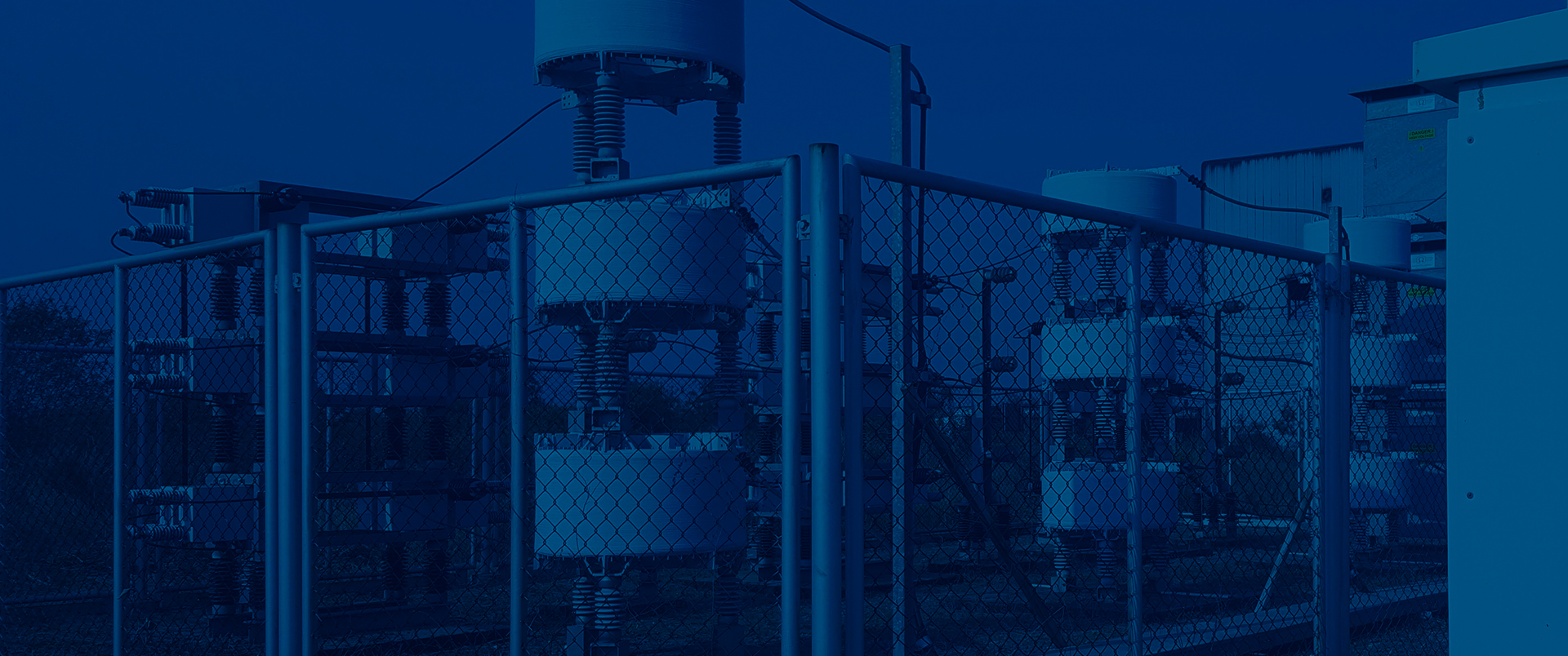 Electrical substation equipment including transformers, insulators, and switchgear behind chain-link fencing, with a dark blue color overlay